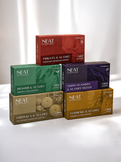 Six boxed herbal ghee products from NEAT Everyday arranged in a pyramid-like stack on a white studio background. The boxes are in different colors: red, green, dark purple, brown, and gold, each labeled with a different ghee type including Trikatu &amp; A2 Ghee, Brahmī &amp; A2 Ghee, Ashwagandha &amp; A2 Ghee, Triphala &amp; A2 Ghee, and Turmeric &amp; A2 Ghee.